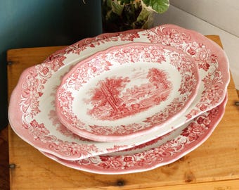 May include: Three oval serving platters with a pink floral and landscape design. The platters are stacked on top of each other, with the largest one on the bottom and the smallest on top. The platters have a scalloped edge.