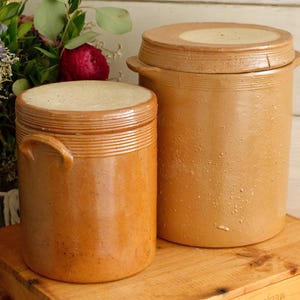 May include: Two antique, cylindrical stoneware crocks with lids. The crocks are a warm, earthy brown color with a textured surface. One crock has a handle. The lids are flat and fit snugly on top. The crocks are on a wooden surface.