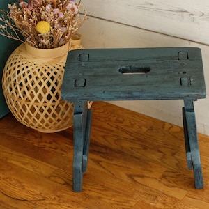 Puede incluir: Un pequeño taburete de madera azul-verdoso con una parte superior rectangular y patas plegables. El taburete tiene un acabado envejecido y un asa recortada en la parte superior. Una cesta tejida con flores secas está cerca.