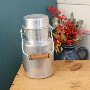 Puede incluir: Una jarra de leche de aluminio plateado con un asa de madera. La jarra tiene una tapa y un asa de transporte. La jarra está sobre una superficie de madera.