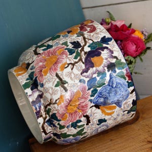 May include: A ceramic planter featuring a white background with a colourful floral pattern of pink, purple, and yellow flowers, green leaves, and a raised blue ram head relief on the side. The cylindrical vessel has a wide opening and a smooth finish.