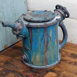 Pode incluir: Uma chaleira de cerâmica azul e marrom com tampa e alça de metal. A chaleira tem um visual rústico e industrial.