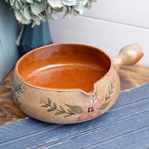 May include: A brown ceramic pot with a handle and a floral design. The pot has a brown glaze with a light brown base and a floral design in shades of pink, green, and brown.