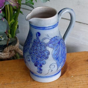 May include: A light blue ceramic pitcher with a handle and spout. The pitcher features a detailed blue grape and leaf design. The pitcher is approximately 9 inches tall.