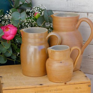 Puede incluir: Tres jarras de cerámica marrón de diferentes tamaños con un glaseado gris claro. Las jarras están dispuestas sobre una caja de madera con un fondo blanco.