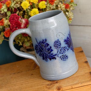 May include: A light gray ceramic beer mug with a single handle, featuring a prominent embossed design of dark blue hop cones and leaves wrapping around the side. The surface has a smooth, glazed finish with a subtle speckled texture throughout.