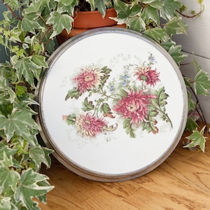 May include: A white ceramic plate with a silver rim features a floral design of pink and white chrysanthemums with green leaves. The plate is on a wooden surface.
