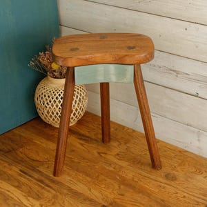 Puede incluir: Taburete de madera con diseño de tres patas. El asiento es de color marrón claro con tres pequeñas incrustaciones decorativas. Una sección pintada de azul claro es visible debajo del asiento. El taburete está sobre un suelo de madera.