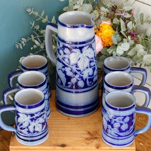 May include: A ceramic beverage set consisting of one tall pitcher and six matching mugs. Each piece features a two-tone design with a light gray base and deep blue accents, decorated with embossed relief patterns of blackberry leaves and fruit.