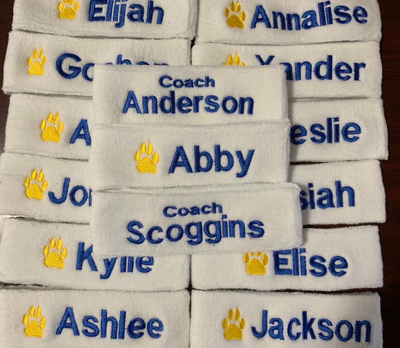 Puede incluir: Bandas blancas de rizo bordadas con nombres en hilo azul. Cada banda presenta una huella de pata amarilla y nombres personalizados como "Elijah", "Abby" y "Coach Anderson".