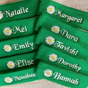 Puede incluir: Bandas verdes de rizo bordadas con nombres en escritura blanca y una flor de margarita. Los nombres incluyen Natalie, Margaret, Mei, Dara, Emily, Tavishi, Elise, Dorothy, Noushee y Hannah. Las bandas son de un verde vibrante.