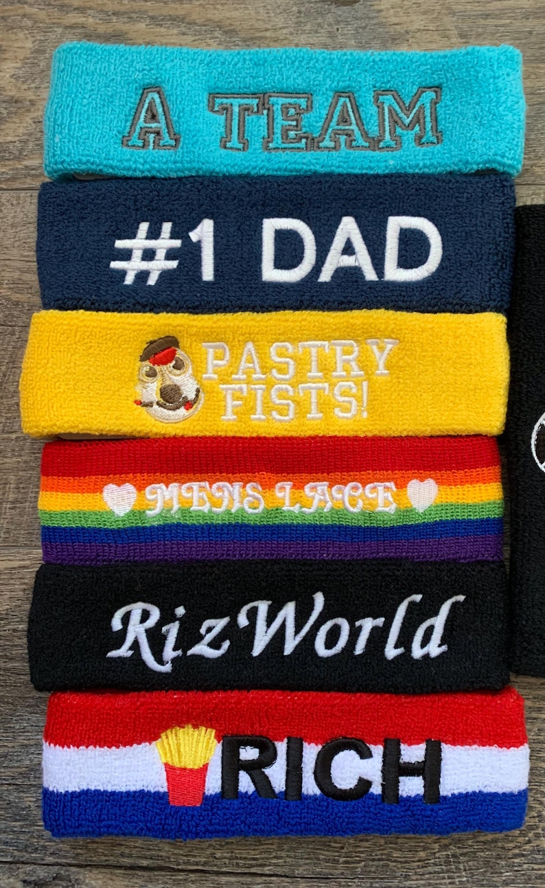 May include: A stack of colorful terry cloth headbands. The headbands are teal, navy, yellow, rainbow, black, and red, white, and blue. Each headband has embroidered text: "A TEAM", "#1 DAD", "PASTRY FISTS!", "MENS LACE", "RizWorld", and "RICH".