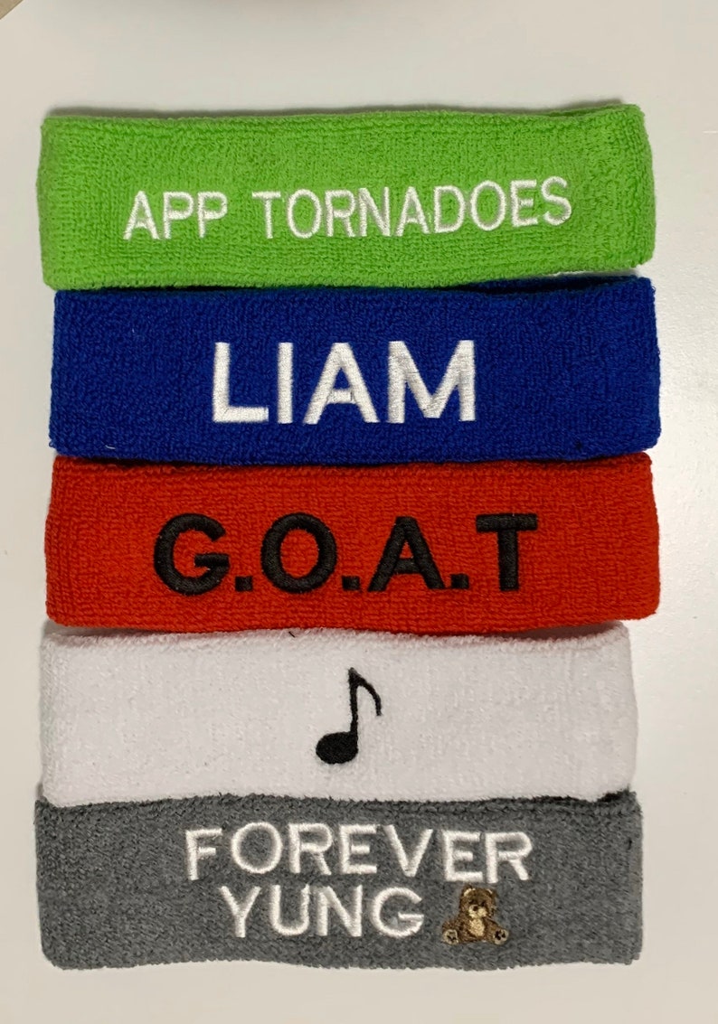 May include: A stack of five terry cloth headbands in various colors. The green headband has "APP TORNADOES" embroidered in white. The blue headband has "LIAM" embroidered in white. The red headband has "G.O.A.T" embroidered in black. The white headband has a black musical note. The gray headband has "FOREVER YUNG" embroidered in white with a small teddy bear.