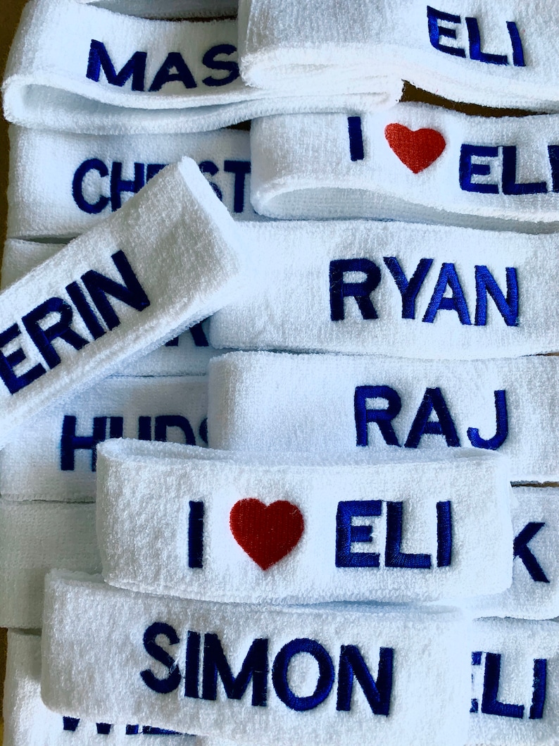 Puede incluir: Una pila de cintas para la cabeza blancas de rizo con texto bordado en azul. Algunas cintas muestran nombres como "RYAN" y "SIMON". Una cinta tiene un coraz&oacute;n rojo y el texto "I ❤️ ELI."