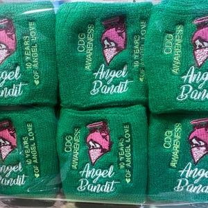 Puede incluir: Un conjunto de seis mu&ntilde;equeras verdes de rizo. Cada banda presenta las palabras "CDG Awareness", "Angel Bandit" y "10 Years of Angel Love" en escritura blanca. Tambi&eacute;n hay un gr&aacute;fico rosa y blanco bordado.