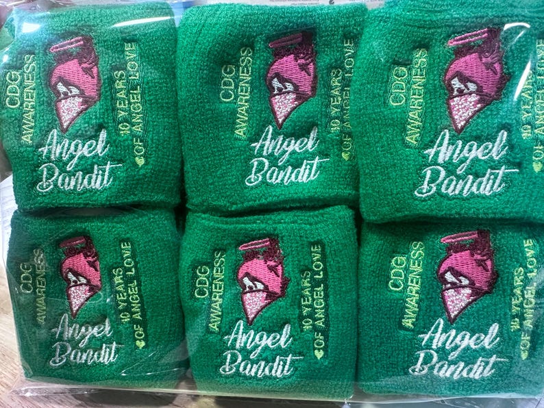 Puede incluir: Un conjunto de seis mu&ntilde;equeras verdes de rizo. Cada banda presenta las palabras "CDG Awareness", "Angel Bandit" y "10 Years of Angel Love" en escritura blanca. Tambi&eacute;n hay un gr&aacute;fico rosa y blanco bordado.