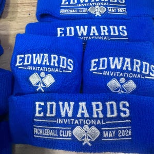 Puede incluir: Varias mu&ntilde;equeras azules de rizo bordadas con "EDWARDS INVITATIONAL" y "PICKLEBALL CLUB MAY 2026" en blanco. Las mu&ntilde;equeras presentan un logotipo de raqueta y pelota de pickleball. El texto es claro.