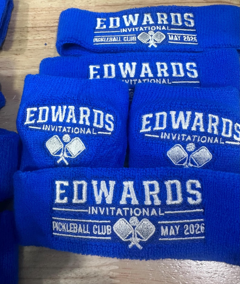 Puede incluir: Varias mu&ntilde;equeras azules de rizo bordadas con "EDWARDS INVITATIONAL" y "PICKLEBALL CLUB MAY 2026" en blanco. Las mu&ntilde;equeras presentan un logotipo de raqueta y pelota de pickleball. El texto es claro.
