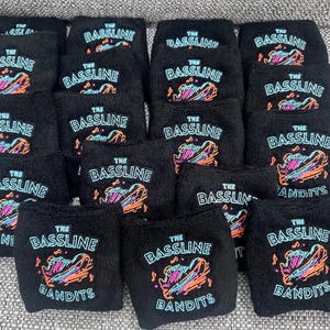 Puede incluir: Una colecci&oacute;n de mu&ntilde;equeras negras con el texto "THE BASSLINE BANDITS" en blanco y un gr&aacute;fico colorido. El gr&aacute;fico presenta un instrumento musical estilizado con acentos naranjas, azules y rosas. Las mu&ntilde;equeras est&aacute;n hechas de un material suave y absorbente.