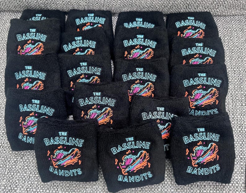 Puede incluir: Una colecci&oacute;n de mu&ntilde;equeras negras con el texto "THE BASSLINE BANDITS" en blanco y un gr&aacute;fico colorido. El gr&aacute;fico presenta un instrumento musical estilizado con acentos naranjas, azules y rosas. Las mu&ntilde;equeras est&aacute;n hechas de un material suave y absorbente.
