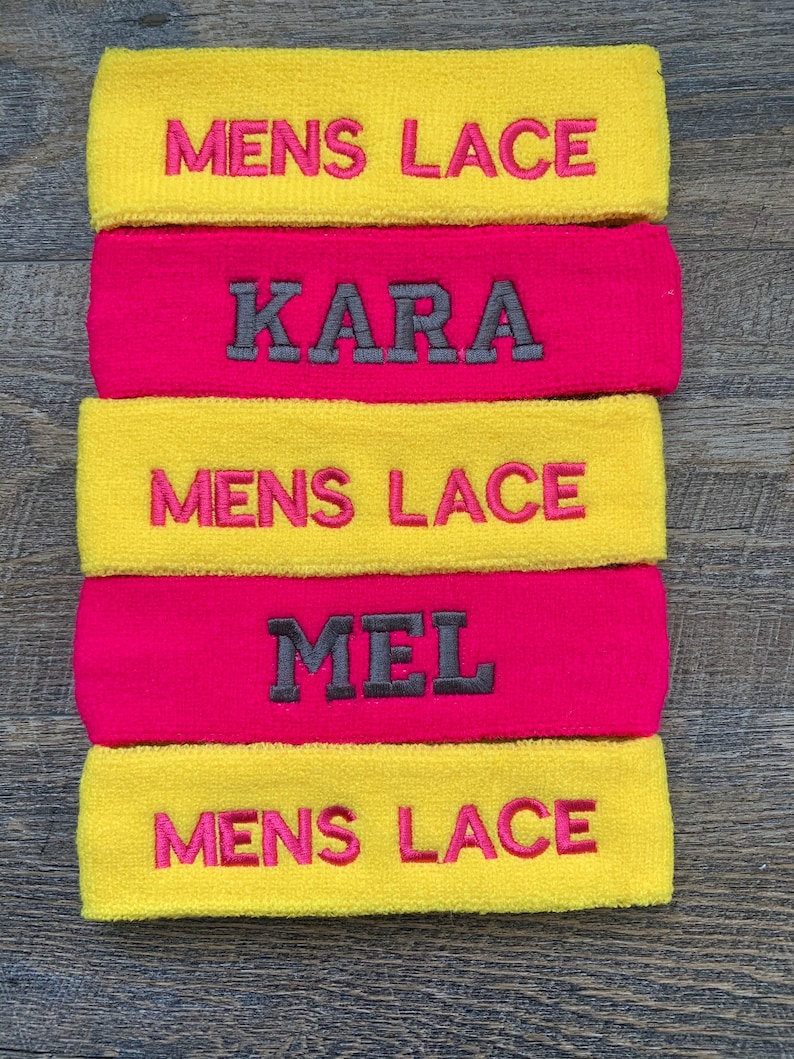 Puede incluir: Una pila de cinco cintas para la cabeza de rizo. Dos son amarillas con "MENS LACE" bordado en rosa. Dos son de color rosa intenso con nombres bordados en gris. Una es rosa intenso con "MEL" bordado en gris.