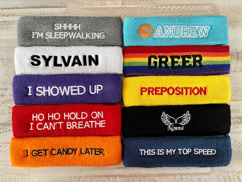 May include: A collection of colorful headbands with embroidered text. The headbands are in various colors, including gray, white, purple, red, orange, blue, yellow, black, and a rainbow stripe. Text includes phrases and names.