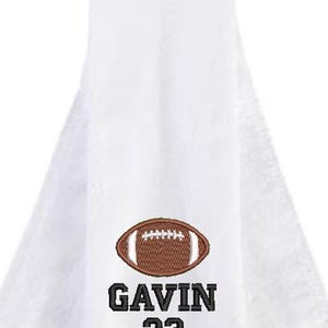May include: White hand towel with an embroidered brown football design. The name "GAVIN" and the number "23" are embroidered below the football in black.