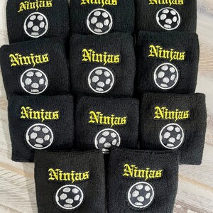 Puede incluir: Un conjunto de mu&ntilde;equeras negras bordadas con la palabra "Ninjas" en letras g&oacute;ticas amarillas y un dise&ntilde;o de bal&oacute;n de f&uacute;tbol blanco y negro. Estas mu&ntilde;equeras est&aacute;n dise&ntilde;adas para deportes o actividades atl&eacute;ticas.