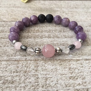 May include: A beaded bracelet featuring a combination of purple, pink, black, and clear beads. The bracelet includes a large pink bead, smaller clear and black beads, and purple beads. The beads are strung on a stretchy cord.