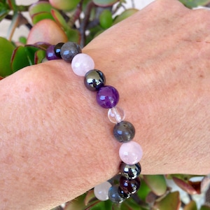 May include: A bracelet made of various gemstones, including amethyst, rose quartz, hematite, and labradorite. The bracelet is worn on a person's wrist.