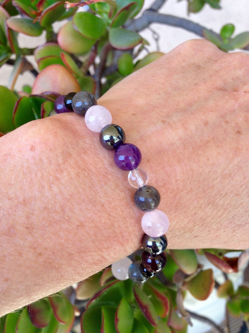 May include: A bracelet made of various gemstones, including amethyst, rose quartz, hematite, and labradorite. The bracelet is worn on a person's wrist.