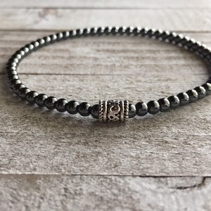 May include: A dark gray beaded bracelet with a decorative silver-colored cylindrical bead. The bracelet is made of small, round beads and is displayed on a light gray wooden surface. The bracelet is a simple, elegant accessory.