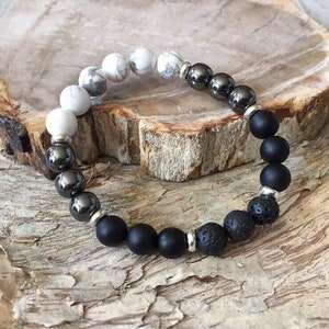May include: A beaded bracelet featuring a mix of white, gray, black, and silver beads. The bracelet includes white marble-like beads, shiny gray beads, matte black beads, and textured lava beads, strung together with silver-colored spacers.