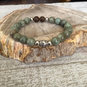 May include: A beaded bracelet featuring green and brown stone beads. The bracelet has a silver-colored decorative bead in the center. The beads are strung on an elastic band. The bracelet is displayed on a wooden surface.