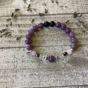 May include: A purple, lavender, and pink gemstone bracelet with a black lava bead and silver accents. The bracelet is on a light gray wooden surface.