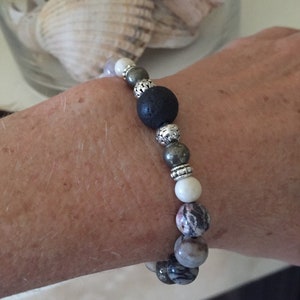 May include: A beaded bracelet featuring a variety of natural stone beads in shades of black, gray, white, and pink. The bracelet includes a large, dark gray lava stone bead and decorative silver-colored spacers. The bracelet is shown on a wrist.