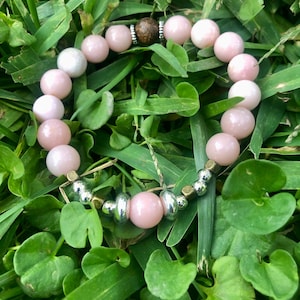 May include: A pink and white beaded bracelet with silver and gold accents. The bracelet is lying on a bed of green grass.
