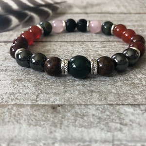 May include: A beaded bracelet featuring a variety of natural stone beads in shades of red, brown, green, black, and pink. The bracelet includes a mix of polished and matte beads, strung together with a stretch cord. Silver-colored spacers are also included.