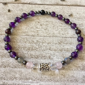 May include: A beaded bracelet featuring amethyst, hematite, rose quartz, and labradorite stones. The bracelet includes a silver-colored cylindrical bead with an ornate design. The beads are strung on an elastic cord.