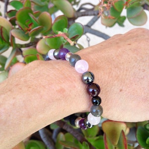 May include: A bracelet made of various gemstones, including amethyst, rose quartz, hematite, and garnet. The bracelet is on a person's wrist.