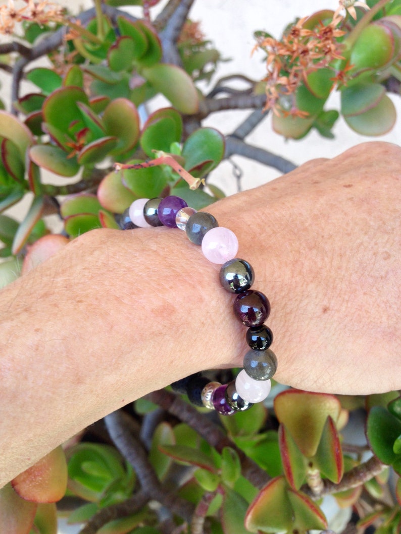 May include: A bracelet made of various gemstones, including amethyst, rose quartz, hematite, and garnet. The bracelet is on a person's wrist.