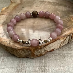 Puede incluir: Un brazalete de piedras preciosas rosas con cuentas de plata y bronce. El brazalete está sobre una superficie de madera.
