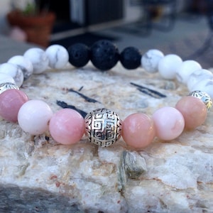 May include: A bracelet made with white, pink, and black beads. The bracelet has a silver charm with a geometric design.