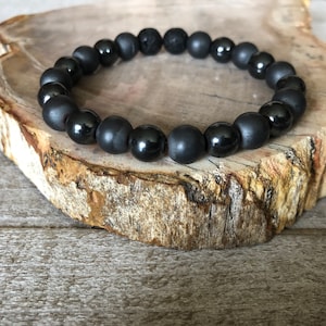 May include: A black beaded bracelet featuring a mix of matte and glossy round beads. The bracelet is made of various black stones, including what appears to be lava rock. The bracelet is displayed on a piece of wood with a natural, rustic appearance.