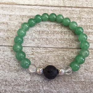 May include: A stretch bracelet featuring smooth, round, translucent green beads, a single porous black lava bead at the center, two clear crystal beads, and two small silver-toned metal spacer beads with a decorative etched pattern.