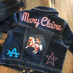 May include: A dark blue denim jacket with the name "Mary Claire" embroidered in pink on the back. The jacket features a colorful embroidered design of a cowgirl on a horse, along with a blue arrow and a pink star.