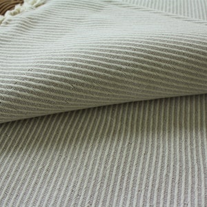 May include: A light beige fabric featuring a subtle herringbone weave pattern. The textile is folded over to reveal the texture of the weave and has a frayed edge with loose threads visible along the top left corner of the material.