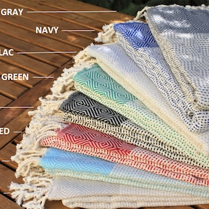 May include: A stack of eight Turkish towels in various colors, including beige, blue, red, black, green, lilac, navy, and gray. The towels are folded and arranged in a pyramid shape, with the beige towel at the bottom and the gray towel at the top.