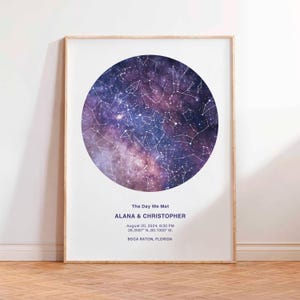 Peut inclure: Une affiche encadrée avec une carte du ciel circulaire, aux tons violets et bleus. Le texte "The Day We Met" surmonte les noms ALANA & CHRISTOPHER, avec une date, une heure et un lieu. Le cadre est en bois clair.