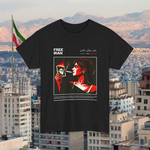 May include: Black t-shirt featuring the text "FREE IRAN" and an illustration of a woman smoking a cigarette while holding a burning photo. The image also includes text in Farsi and English, with a cityscape and a flag in the background.